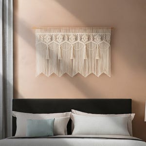 May include: A cream-colored macrame wall hanging with a geometric design and fringe. The wall decor is mounted on a wooden dowel and hangs above a bed with pillows. The macrame has a scalloped bottom edge.