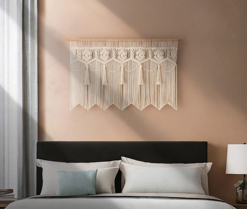 May include: A cream-colored macrame wall hanging with a geometric design and fringe. The wall decor is mounted on a wooden dowel and hangs above a bed with pillows. The macrame has a scalloped bottom edge.