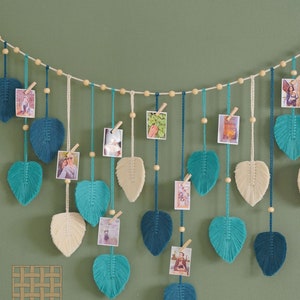 May include: A string of macrame leaf-shaped wall decor with wooden beads and clips for hanging photos. The leaves are in shades of teal, blue, and cream.