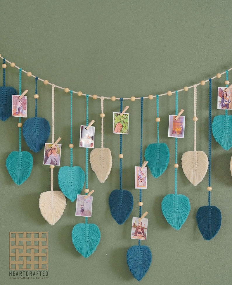 May include: A string of macrame leaf-shaped wall decor with wooden beads and clips for hanging photos. The leaves are in shades of teal, blue, and cream.