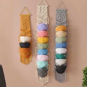May include: Three vertical macrame wall organizers made of knotted cord, each featuring multiple pockets holding various baseball caps. The left organizer is mustard yellow, the center is cream, and the right is light gray, all with fringe.