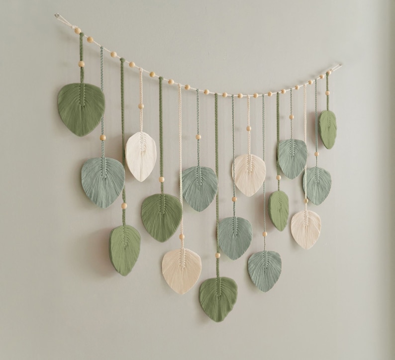 May include: A wall hanging made of macrame with green and cream leaves. The leaves are strung on a string with wooden beads.