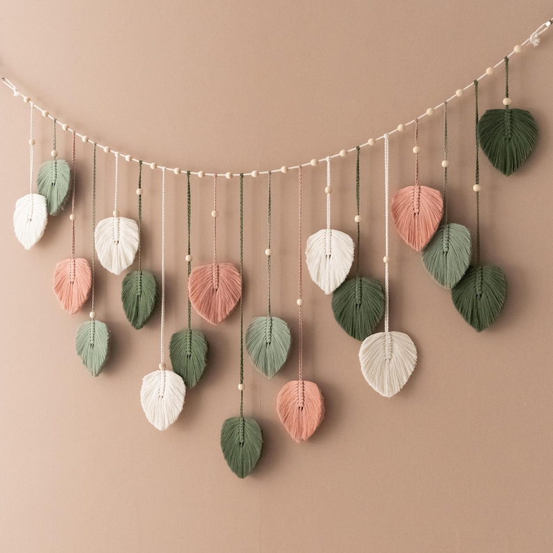 May include: A garland of macrame leaves in shades of green, white, and pink, strung on a white cord with wooden beads.