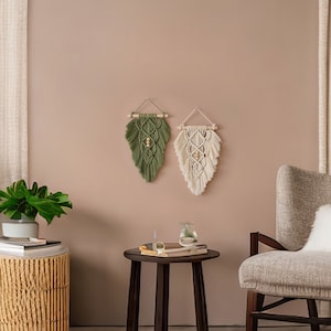 May include: Two macrame wall hangings, one green and one white, with wooden dowels and beads, hanging on a light brown wall. A brown side table with a book and a glass vase sits in front of a beige armchair.