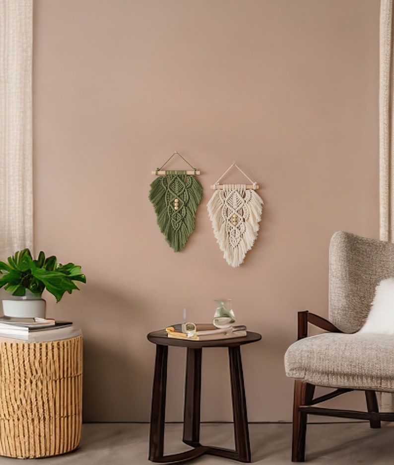 May include: Two macrame wall hangings, one green and one white, with wooden dowels and beads, hanging on a light brown wall. A brown side table with a book and a glass vase sits in front of a beige armchair.