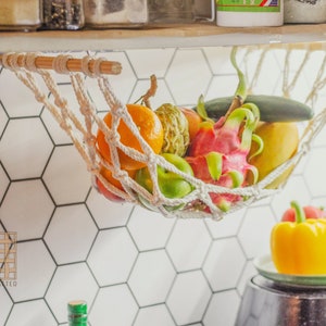May include: A white rope hammock hanging from a wooden shelf, filled with fresh fruit, including a dragon fruit, a yellow mango, a green cucumber, and a few oranges. The hammock is hanging over a white hexagonal tile wall.