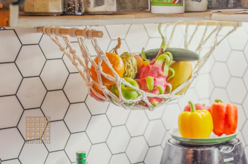 May include: A white rope hammock hanging from a wooden shelf, filled with fresh fruit, including a dragon fruit, a yellow mango, a green cucumber, and a few oranges. The hammock is hanging over a white hexagonal tile wall.