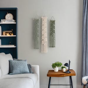May include: Three macrame wall hangings with a leaf pattern. The hangings are in shades of green and cream. The hangings are displayed on a white wall in a living room setting.