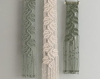 Puede incluir: Tres tapices de macramé verticales colgados de varillas de madera. Dos tapices son de color verde salvia y el central es de color blanco roto. Cada uno presenta un patrón de nudos en forma de hoja con flecos y pequeños nudos.