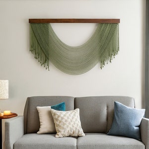 May include: A gray sofa with three pillows, one blue, one white, and one patterned with beige and white. A green macrame wall hanging with a wooden frame is above the sofa.