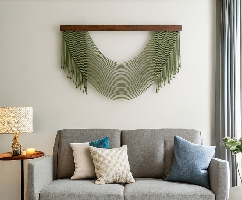 May include: A gray sofa with three pillows, one blue, one white, and one patterned with beige and white. A green macrame wall hanging with a wooden frame is above the sofa.