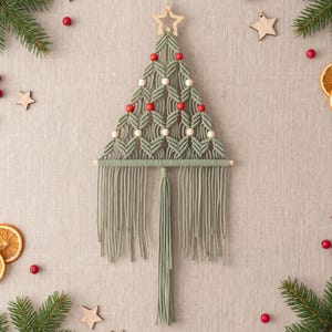 Op de afbeelding: Een wandhanger in de vorm van een kerstboom gemaakt van saliegroen macramékoord. Het ontwerp is voorzien van een houten ster aan de bovenkant, afwisselende rijen rode en natuurlijke houten kralen en lange franjes die aan een stok hangen.