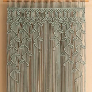 Pode incluir: Uma tapeçaria de macramé de parede com um design de folhas em uma cor verde salvia clara. A tapeçaria é feita com uma série de nós e cordas, criando um padrão texturizado e intrincado.