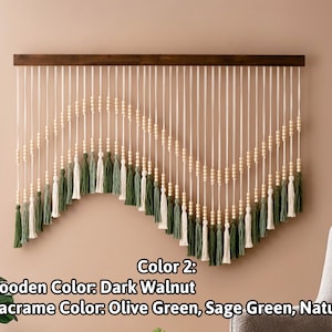 May include: A wall hanging made of macrame with wooden beads and tassels in olive green, sage green, and natural colors. The macrame is attached to a dark walnut wood bar.
