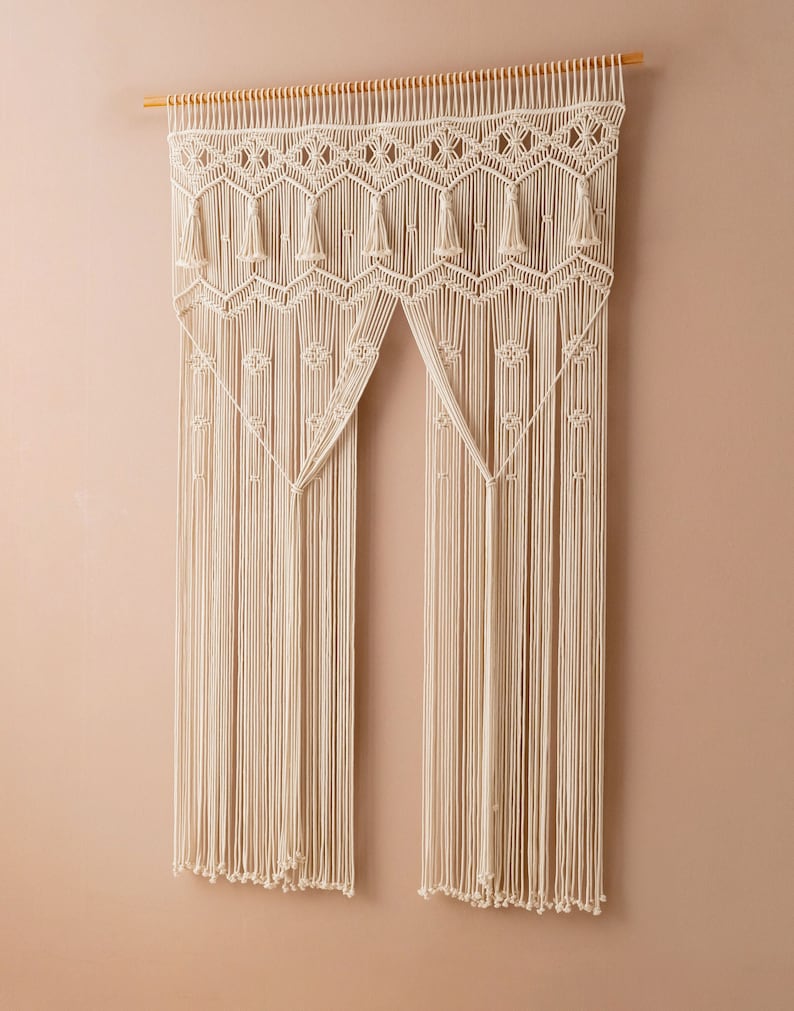 May include: A white macrame wall hanging with a geometric pattern and tassels. The hanging is made of cotton cord and is attached to a wooden dowel.