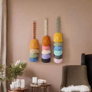 K&ouml;nnte beinhalten: Eine Wand mit neun Baseballkappen, die an Makramee-Wandhaltern h&auml;ngen. Die Kappen sind in verschiedenen Farben, darunter Gelb, Orange, Rosa, Blau und Gr&uuml;n.
