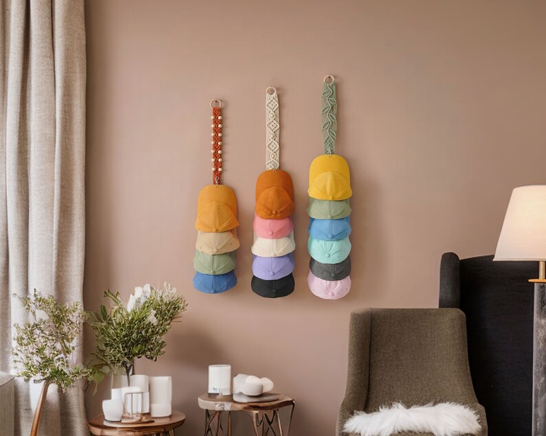 K&ouml;nnte beinhalten: Eine Wand mit neun Baseballkappen, die an Makramee-Wandhaltern h&auml;ngen. Die Kappen sind in verschiedenen Farben, darunter Gelb, Orange, Rosa, Blau und Gr&uuml;n.