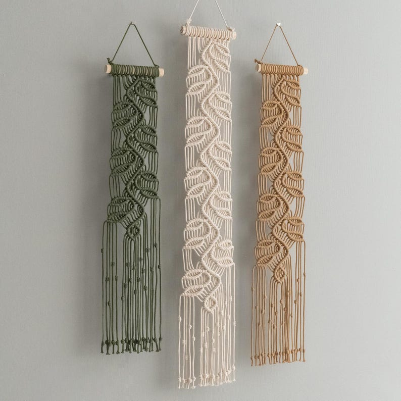 May include: Three macrame wall hangings suspended from wooden dowels with string loops. Each piece features a vertical leaf-like pattern created with knotted cord in olive green, cream, and tan. The bottom edges consist of long, loose fringe.
