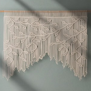 May include: A white macrame wall hanging with a leaf design. The hanging has a fringe border and is made of cotton cord.