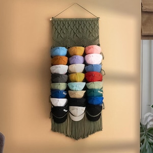 May include: A green macrame wall hanging with a fringe border. The wall hanging has 20 slots for storing baseball caps. The caps are in a variety of colours, including blue, red, yellow, and white.