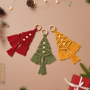 Puede incluir: Tres adornos de &aacute;rbol de Navidad de macram&eacute; hechos a mano en rojo, verde y amarillo con cuentas de madera y borlas. Los adornos cuelgan de anillos de madera.