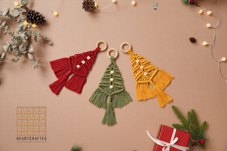 Puede incluir: Tres adornos de &aacute;rbol de Navidad de macram&eacute; hechos a mano en rojo, verde y amarillo con cuentas de madera y borlas. Los adornos cuelgan de anillos de madera.