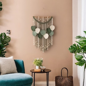 Op de afbeelding: Een macram&eacute; wandhanger met groene en witte bladeren, hangend aan een lichtbruine muur. De wandhanger is gemaakt van garen en kralen. Er is een groene plant in een pot aan de linkerkant van de afbeelding en een groene plant in een pot aan de rechterkant van de afbeelding.