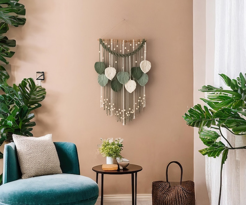 Op de afbeelding: Een macram&eacute; wandhanger met groene en witte bladeren, hangend aan een lichtbruine muur. De wandhanger is gemaakt van garen en kralen. Er is een groene plant in een pot aan de linkerkant van de afbeelding en een groene plant in een pot aan de rechterkant van de afbeelding.