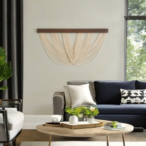 May include: A wall hanging featuring a horizontal dark brown wooden bar with multiple off-white cotton cords draped in a series of overlapping, symmetrical U-shaped loops that create a layered, cascading pattern across the wall surface.