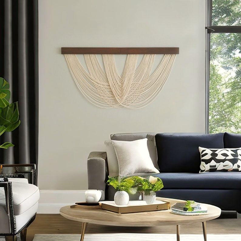 May include: A wall hanging featuring a horizontal dark brown wooden bar with multiple off-white cotton cords draped in a series of overlapping, symmetrical U-shaped loops that create a layered, cascading pattern across the wall surface.