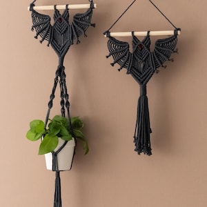 Puede incluir: Dos tapices de pared de macramé negro en forma de murciélagos. Un murciélago cuelga de un colgador de plantas con una pequeña planta verde en una maceta blanca.