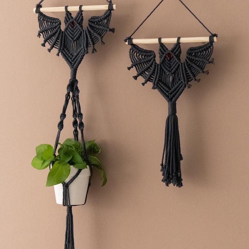Pu&ograve; includere: Due arazzi da parete in macram&eacute; nero a forma di pipistrelli. Un pipistrello &egrave; appeso a un porta piante con una piccola pianta verde in un vaso bianco.