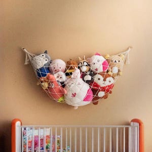 Könnte beinhalten: Eine weiße Hängematte im Makramee-Stil, gefüllt mit verschiedenen Plüschtieren, darunter ein rosa Seestern, ein grauer Wolf und ein weißes Kaninchen mit rosa Akzenten. Die Hängematte ist an einer beigefarbenen Wand über einem weißen Kinderbett mit orangefarbenen Akzenten befestigt.