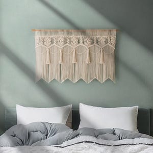 May include: A cream-colored macrame wall hanging with a geometric design and fringe. The wall decor is mounted on a wooden dowel and hangs above a bed with white pillows and a gray comforter. The wall is painted a muted green.
