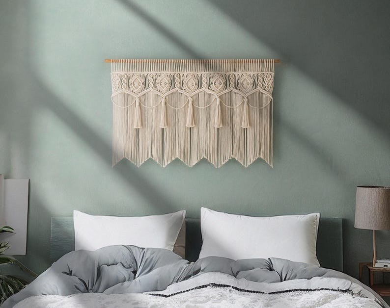 May include: A cream-colored macrame wall hanging with a geometric design and fringe. The wall decor is mounted on a wooden dowel and hangs above a bed with white pillows and a gray comforter. The wall is painted a muted green.