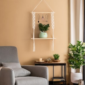 May include: A wooden shelf with macrame hanging on a beige wall. The shelf has a small potted plant and a few pieces of jewellery hanging from it.