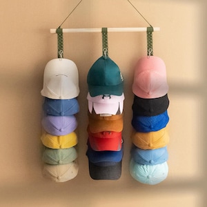 Puede incluir: Un perchero de madera para sombreros con tres filas de gorras de béisbol en varios colores, incluyendo blanco, azul, morado, amarillo, verde, rosa, negro y turquesa. El perchero está suspendido por un cordón verde.