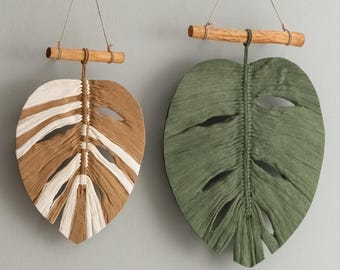 May include: Two macrame wall hangings in the shape of monstera leaves. One is brown and white, the other is green. Both are hanging from wooden dowels.