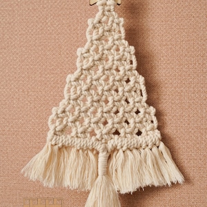 K&ouml;nnte beinhalten: Ein wei&szlig;er Makramee-Weihnachtsbaum-Anh&auml;nger mit einem Holzstern oben. Der Anh&auml;nger hat einen Fransenrand unten und einen langen Quasten, der unten h&auml;ngt.