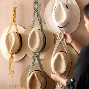 Puede incluir: Tres colgadores de pared de macramé hechos de cuerda de algodón en amarillo mostaza, verde salvia y blanco roto, cada uno con sombreros de paja con bandas negras. Los colgadores presentan nudos complejos y varillas de madera. Texto abajo: HEARTCRAFTED.