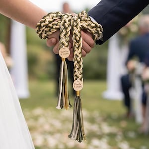 May include: A handfasting cord featuring braided olive green, cream, and gold metallic threads. The cord includes two circular wooden tags engraved with the text 'Camila & Owen' and finishes with long, multi-colored decorative tassels at each end.