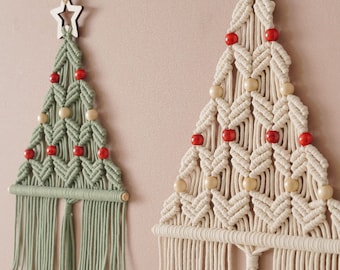 Könnte beinhalten: Zwei Makramee-Wandbehänge in Form von Weihnachtsbäumen. Einer ist salbeigrün, der andere cremeweiß. Beide haben einen Holzstern an der Spitze, rote und naturfarbene Holzperlen im Geflecht sowie lange Fransen am unteren Ende.