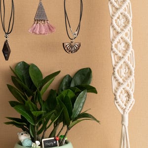 May include: A wooden shelf with a macrame wall hanging, a potted plant, and three pieces of jewellery hanging from hooks. The jewellery includes a necklace with a wooden pendant, a pair of earrings with a pink tassel, and a necklace with a diamond-shaped pendant. The plant is in a light green pot with a small white cat figurine and a sign that says "Happy".