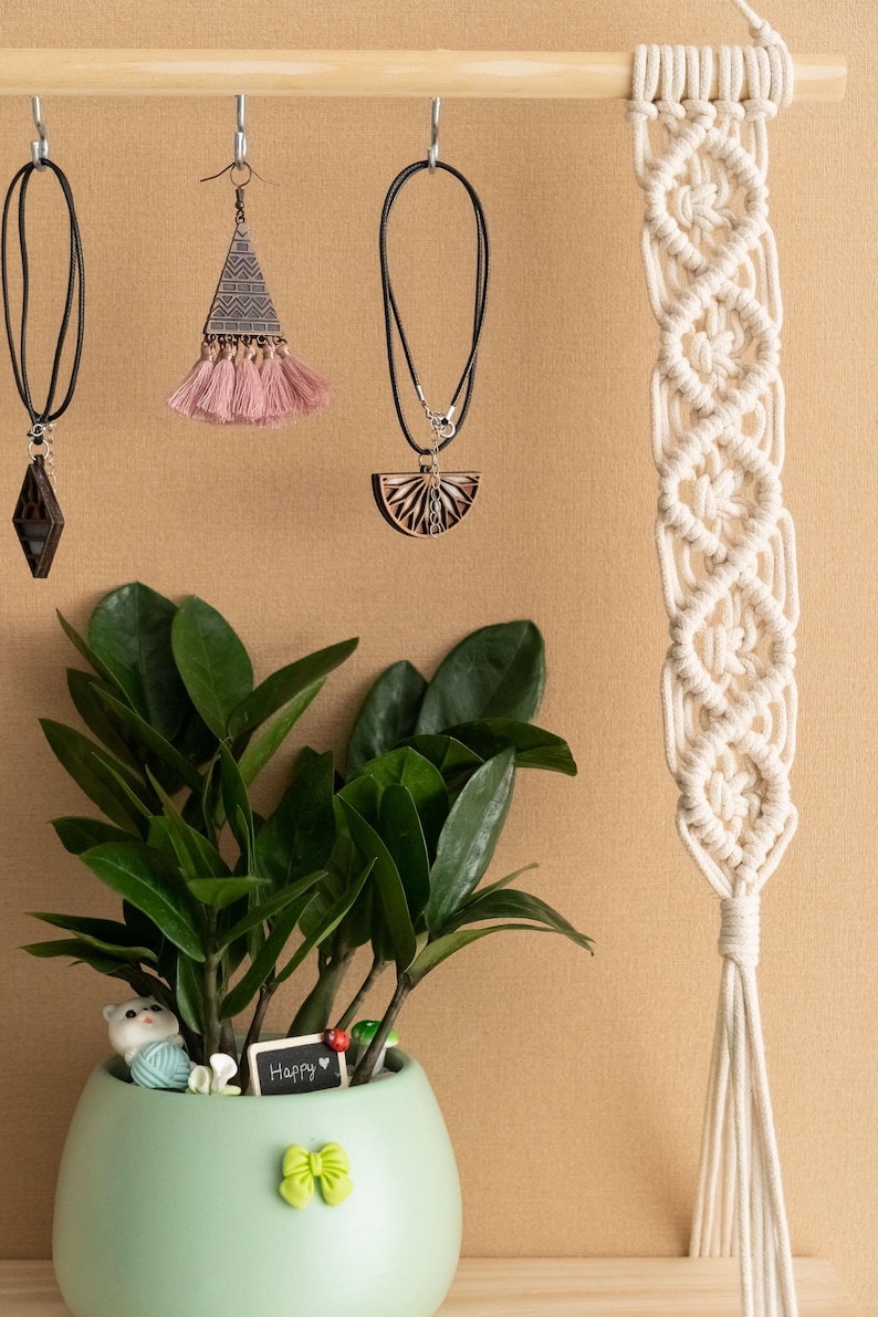 May include: A wooden shelf with a macrame wall hanging, a potted plant, and three pieces of jewellery hanging from hooks. The jewellery includes a necklace with a wooden pendant, a pair of earrings with a pink tassel, and a necklace with a diamond-shaped pendant. The plant is in a light green pot with a small white cat figurine and a sign that says "Happy".
