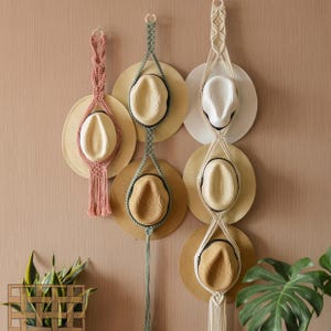 Puede incluir: Tres colgadores de sombreros de macramé en rosa, verde y blanco, cada uno con dos sombreros de paja. Los sombreros tienen un ala beige y una corona color crema. Los colgadores están suspendidos en una pared marrón claro, creando una solución de almacenamiento elegante.