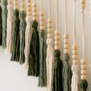 May include: A wall hanging with a series of wooden beads and tassels in shades of green and cream. The tassels are made of yarn and are tied together with string.