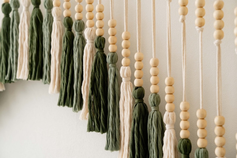 May include: A wall hanging with a series of wooden beads and tassels in shades of green and cream. The tassels are made of yarn and are tied together with string.