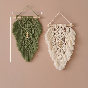 May include: Two macrame wall hangings, one in green and one in cream, with wooden dowels and beads. The hangings are 10.4 inches wide and 13.2 inches high.