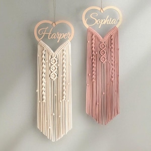 May include: Two wooden heart-shaped wall hangings with macrame fringe. The left one is white and says "Harper" and the right one is pink and says "Sophia".