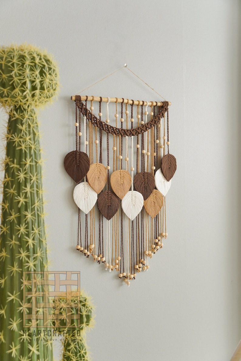 Op de afbeelding: Een bruine, beige en witte macram&eacute; wandhanger met houten kralen en een houten lat aan de bovenkant. De wandhanger heeft de vorm van een traan en heeft een reeks bladeren.